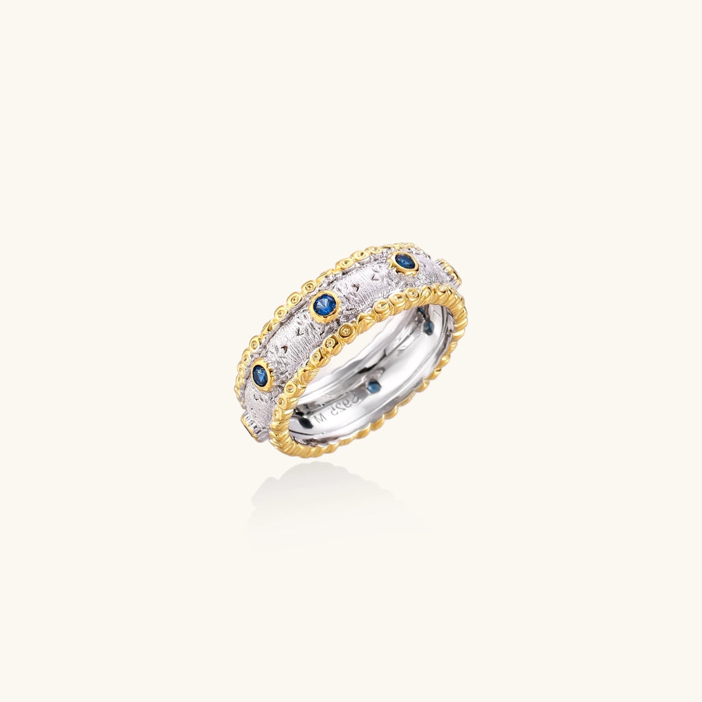 Two-tone ring with brushed silver center, gold beaded edges, and blue zirconia stones on white background.