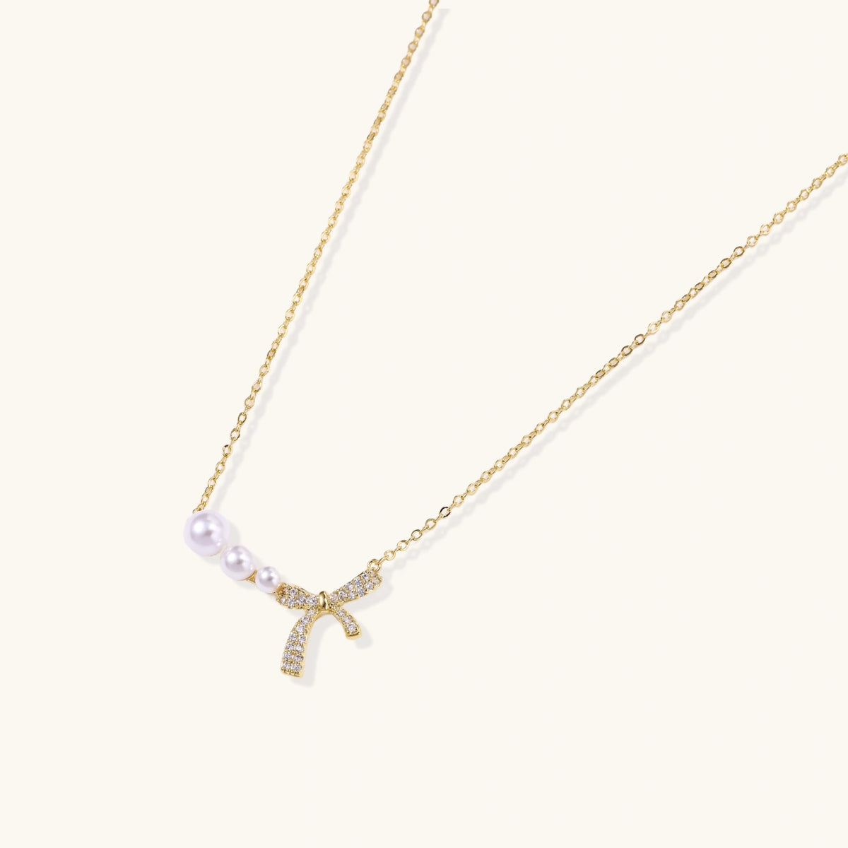 Delicate gold necklace with three white pearls and crystal-embellished bow pendant on cream background