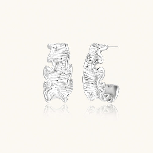 Textured white gold-plated hoop earrings with flowing wave pattern against cream background, showing dimensional rippled surface.