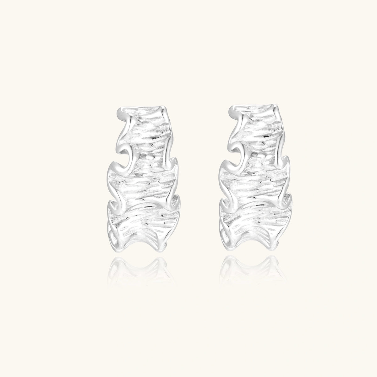 Textured white gold-plated hoop earrings with flowing wave pattern against cream background, showing dimensional rippled surface.