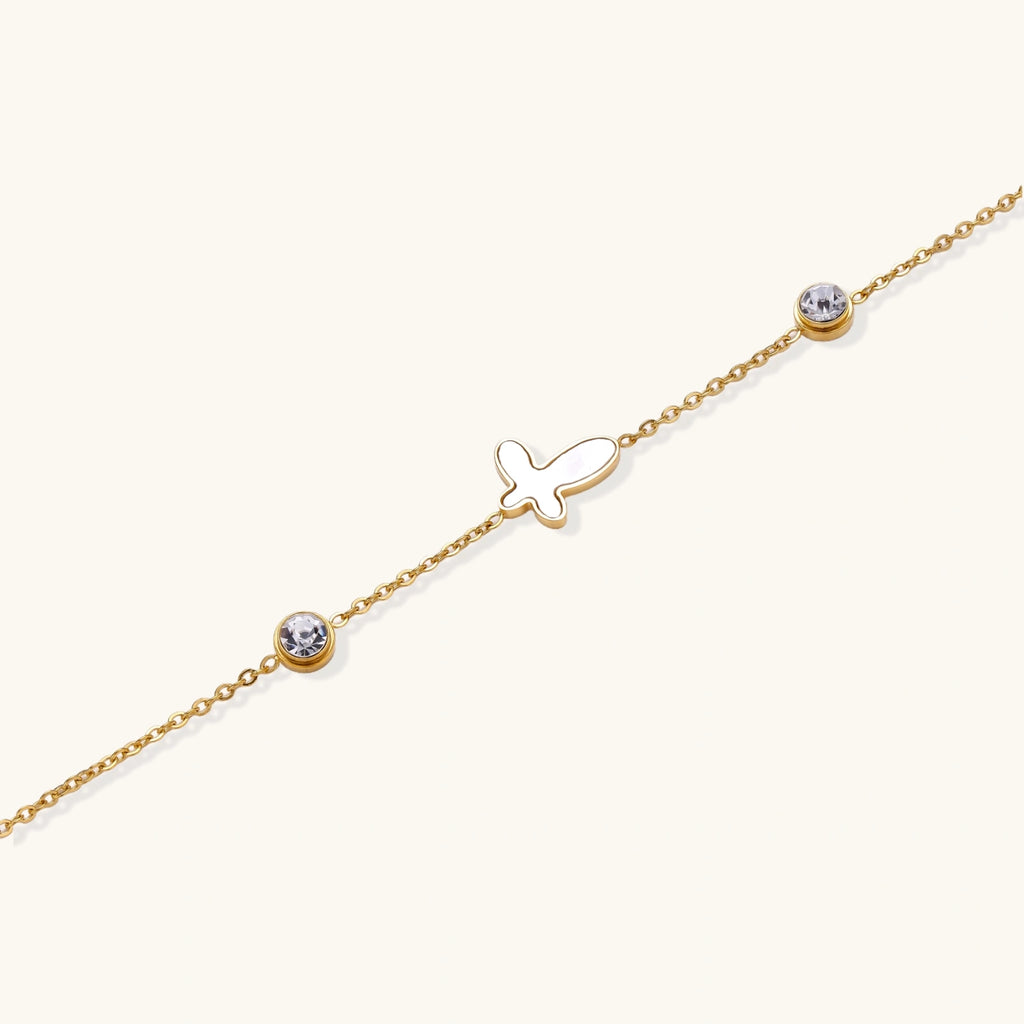 Gold chain bracelet with white mother of pearl butterfly pendant and two bezel-set crystals on cream background